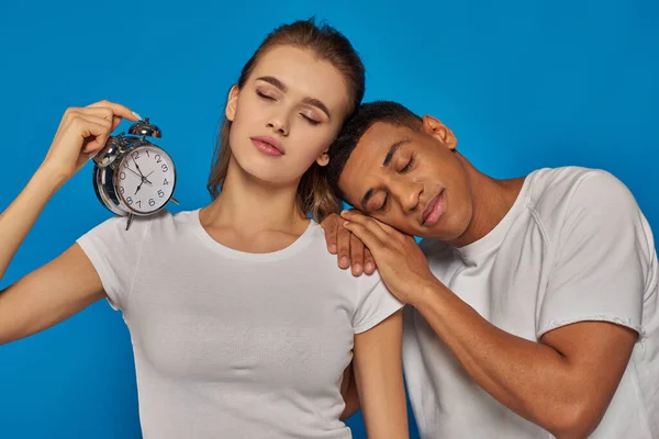 Woman holding alarm clock and sleeping with african american man on blue backdrop, sleepy couple — Stock Photo