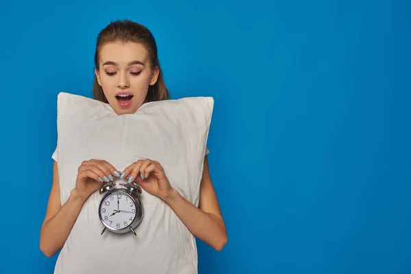 Amazed young woman holding pillow and vintage alarm clock on blue background, early morning — Stock Photo