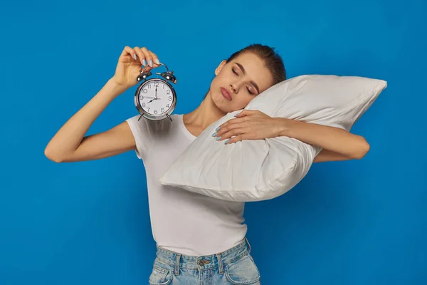Mujer joven disgustado sosteniendo reloj despertador vintage y acostado en la almohada sobre fondo azul, mañana - foto de stock
