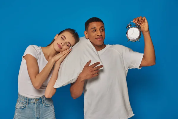 Woman sleeping on pillow near cheerful african american man with alarm clock on blue backdrop — Stock Photo