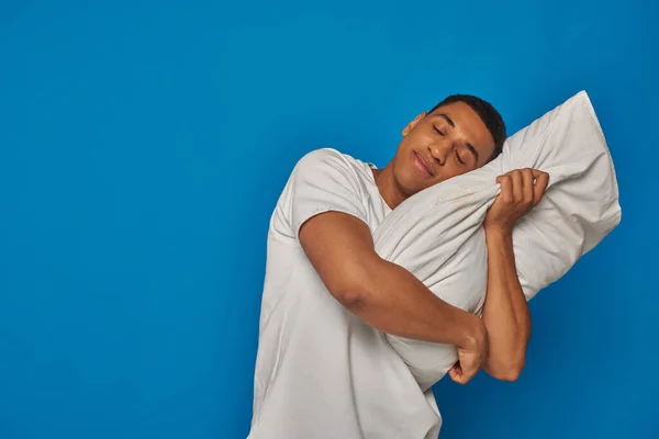 Hombre afroamericano cansado acostado en la almohada y sonriendo sobre fondo azul, persona somnolienta - foto de stock