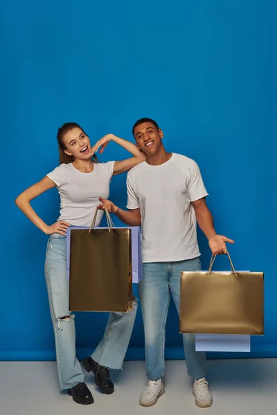 Consumismo, feliz casal interracial segurando sacos de compras e de pé juntos no fundo azul — Fotografia de Stock