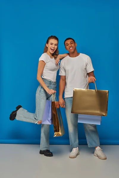 Ventas de vacaciones, feliz pareja interracial sosteniendo bolsas de compras y de pie sobre el telón de fondo azul - foto de stock