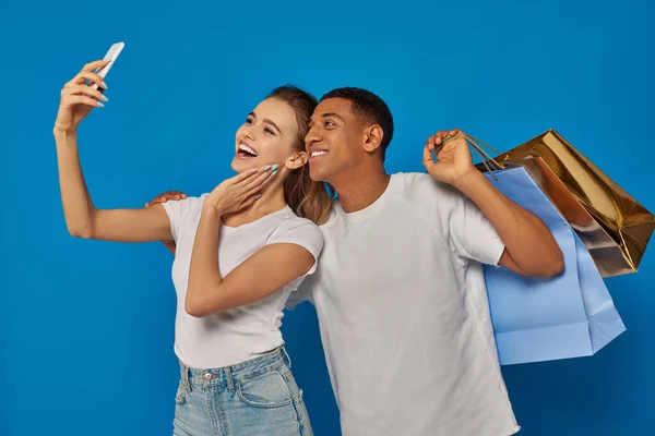 Потребительство, счастливая межрасовая пара, держащая в руках сумки с покупками и делающая селфи на синем рюкзаке — стоковое фото