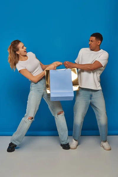 Consumismo, homem interracial e mulher puxando sacos de compras no fundo azul, vendas de férias — Fotografia de Stock