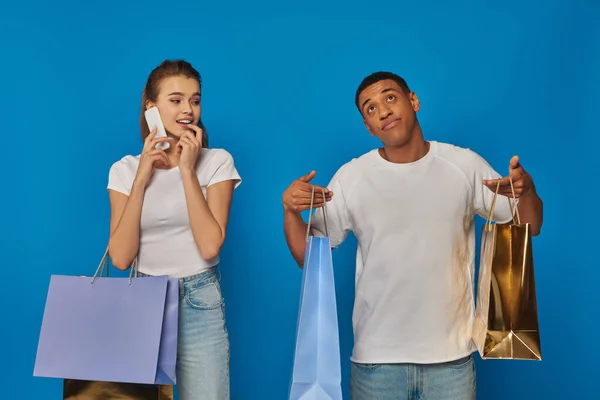 Consumerism, pretty woman having phone call while african american boyfriend holding shopping bags — Stock Photo