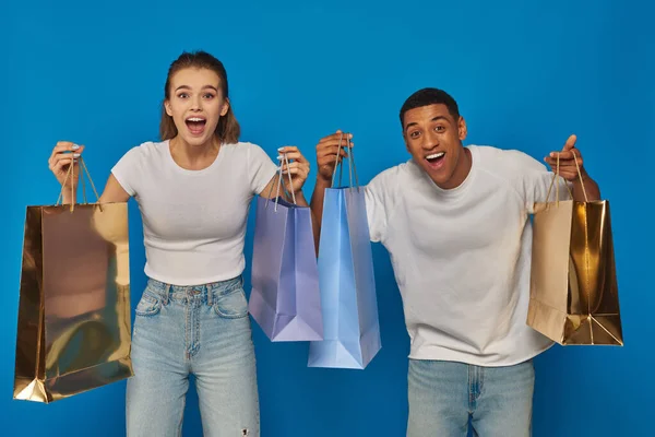 Aufgeregtes gemischtes Paar mit Einkaufstüten und Blick in die Kamera vor blauem Hintergrund, Konsumwahn — Stockfoto