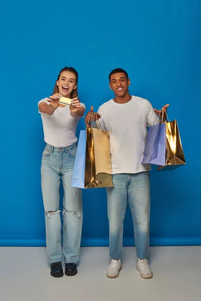 Alegre pareja interracial sosteniendo bolsas de compras y tarjeta de crédito sobre fondo azul, consumismo - foto de stock