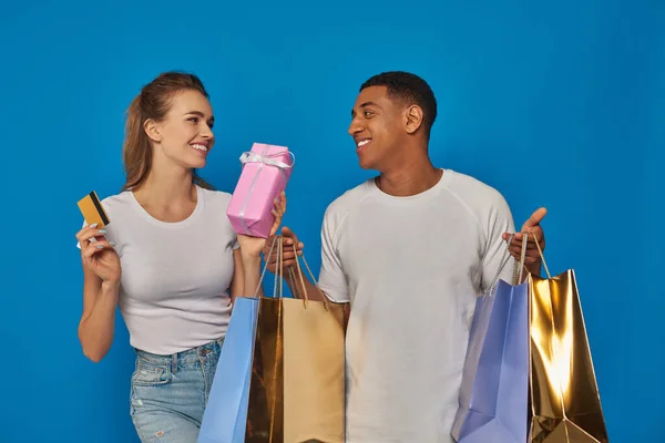 Alegre casal multicultural segurando sacos de compras e cartão de crédito no fundo azul, presente de compra — Fotografia de Stock