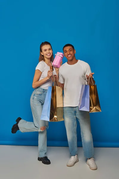 Happy multicultural couple holding shopping bags and credit card on blue backdrop, buying present — Stock Photo