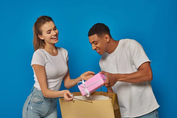 Feliz casal multicultural colocando presente em saco de compras no fundo azul, conceito de consumismo — Fotografia de Stock