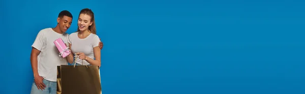 Happy interracial couple buying present together, holding shopping bag on blue backdrop, banner — Stock Photo
