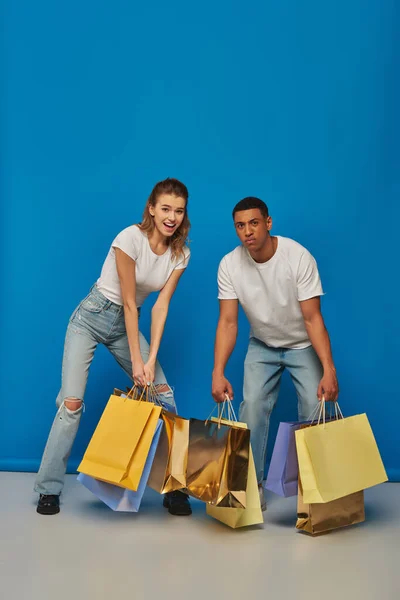 Hombre y mujer multicultural positivo sosteniendo bolsas de compras sobre fondo azul, consumismo y ventas - foto de stock