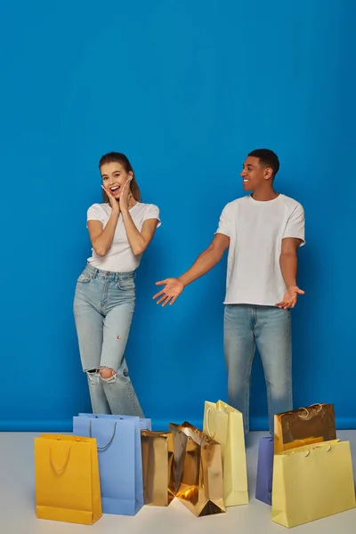 Diversa pareja, hombre afroamericano apuntando a las bolsas de compras cerca de la mujer emocionada en el fondo azul - foto de stock