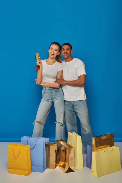 Позитивная мультикультурная пара, стоящая рядом с сумками для покупок и держащая кредитную карту на синем фоне — стоковое фото
