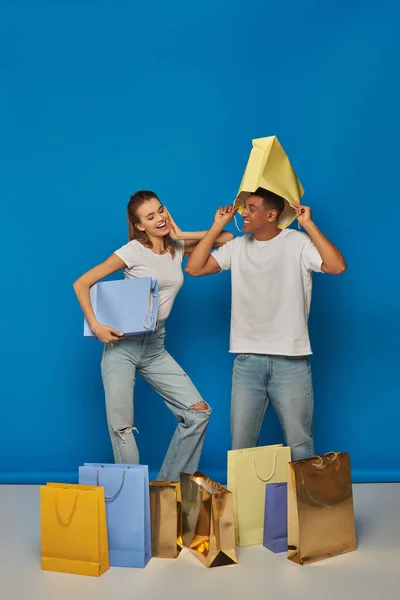 Веселая мультикультурная пара в повседневной одежде, держащая сумки для покупок на синем фоне, розничная радость — стоковое фото