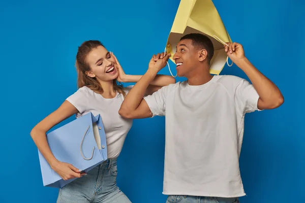 Веселая мультикультурная пара в повседневной одежде, держащая сумки для покупок на синем фоне, покупая веселье — стоковое фото