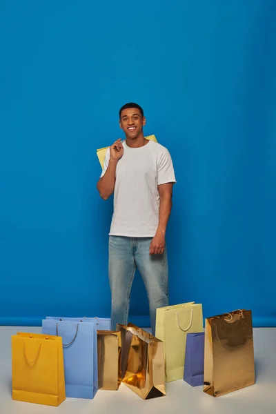 Joyeux homme afro-américain en tenue décontractée tenant des sacs à provisions sur fond bleu, acheter frénésie — Photo de stock