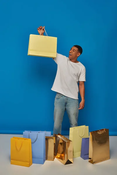 Схвильований афроамериканський чоловік у повсякденному вбранні тримає сумки на синьому фоні, купуючи сплеск — стокове фото