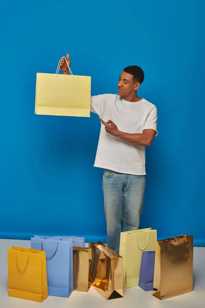 Felice uomo africano americano in abbigliamento casual che punta alla borsa della spesa su sfondo blu, acquistare furia — Foto stock