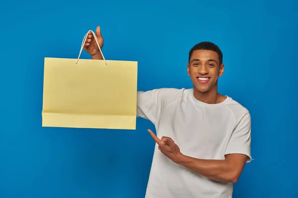 Веселый африканский американец в повседневной одежде, указывающий на сумку с покупками на синем фоне, покупающий — стоковое фото