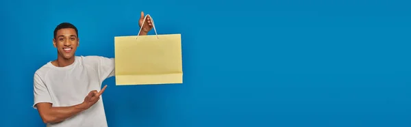 Веселый африканский американец в повседневной одежде, указывающий на сумку для покупок на синем фоне, баннер — стоковое фото