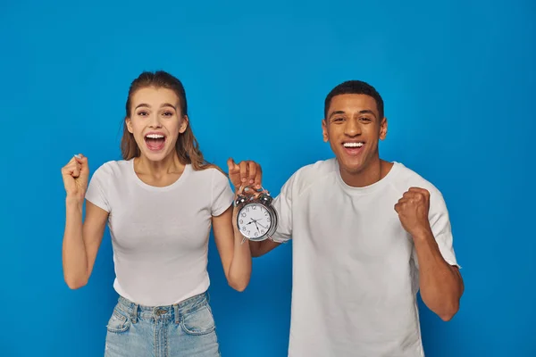 Excited interracial couple with open mouth holding alarm clock on blue background, face expression — Stock Photo