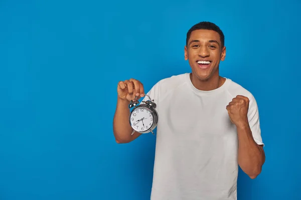 Hombre afroamericano excitado con la boca abierta sosteniendo reloj despertador sobre fondo azul, expresión de la cara - foto de stock
