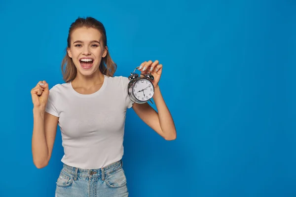 Mujer joven excitada con la boca abierta sosteniendo reloj despertador sobre fondo azul, expresión de la cara - foto de stock