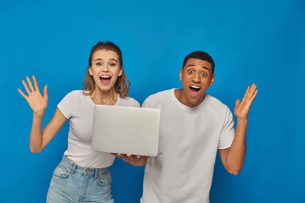 Excité couple multiculturel geste près ordinateur portable sur fond bleu, concept de travail à distance — Photo de stock