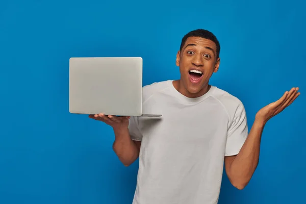 Animado afro-americano homem gestos e segurando laptop em fundo azul, conceito de trabalho remoto — Fotografia de Stock