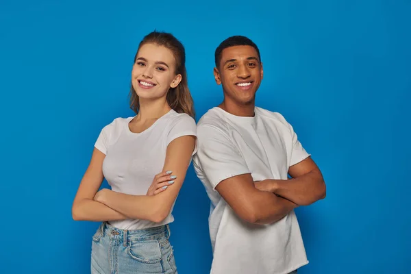 Cheerful multicultural couple standing with folded arms and looking at camera on blue background — Stock Photo