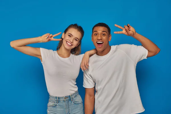 Glückliches multikulturelles Paar zeigt Friedenszeichen und blickt in die Kamera auf blauem Hintergrund, Positivität — Stockfoto