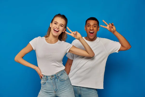 Alegre pareja multicultural mostrando v signo y mirando a la cámara sobre fondo azul, positividad - foto de stock