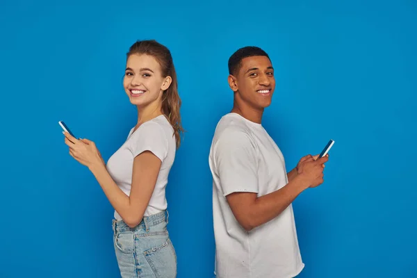 Alegre pareja multicultural de pie espalda con espalda y el uso de teléfonos inteligentes sobre fondo azul - foto de stock