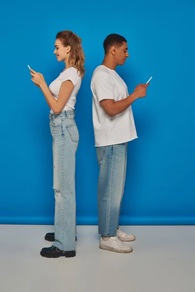 Щаслива мультикультурна пара, що стоїть на спині і використовує смартфони на синьому фоні, вид збоку — стокове фото