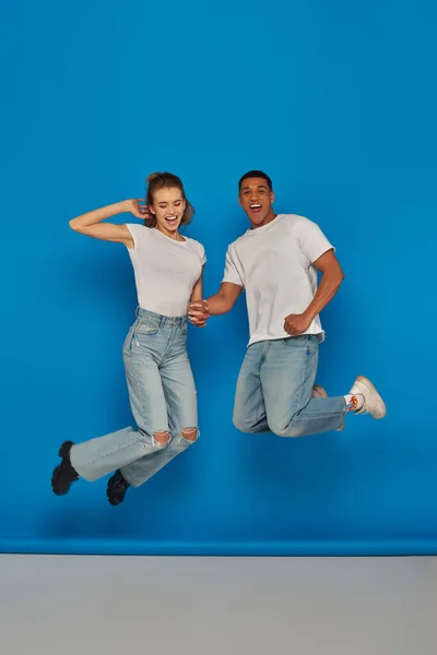 Casal multicultural positivo de mãos dadas e pulando no fundo azul, visão de comprimento total — Fotografia de Stock