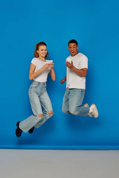 Щаслива мультикультурна пара використовує смартфони і стрибає на синьому фоні, вигляд на повну довжину — стокове фото