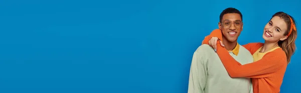 Happy woman in orange cardigan hugging african american man in eyeglasses on blue backdrop, banner — Stock Photo