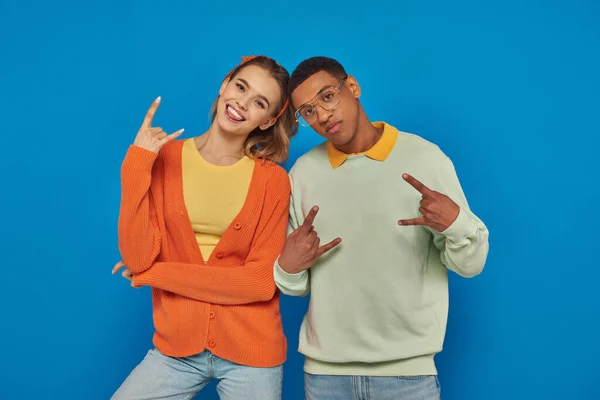 Élégant et jeune couple interracial en tenue décontractée geste sur fond bleu, montrant signe de roche — Photo de stock