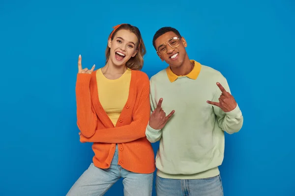 Animado e jovem casal interracial em traje casual gestos no fundo azul, mostrando sinal de rock — Fotografia de Stock