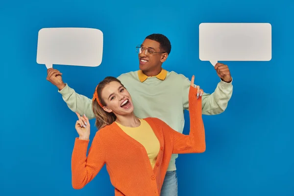 Cheerful diverse couple in smart casual clothes holding blank speech bubbles on blue backdrop — Stock Photo