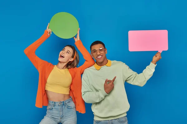 Glückliches interrassisches Paar in smarter Freizeitkleidung mit leeren Sprechblasen auf blauem Hintergrund — Stockfoto