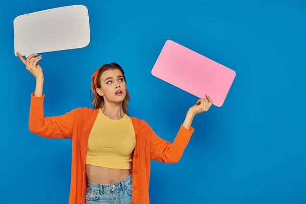 Surprised young woman in casual attire holding speech bubbles on blue background, space for text — Stock Photo