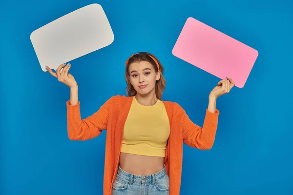 Confused young woman in casual attire holding speech bubbles on blue background, space for text — Stock Photo