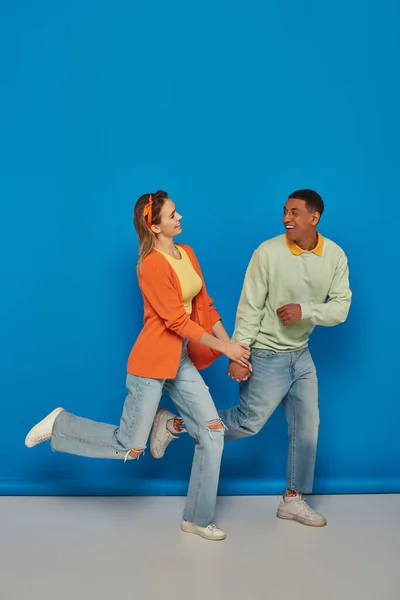 Glückliches multikulturelles Paar in lässiger Kleidung, Händchen haltend und auf blauem Hintergrund laufend, lächelnd — Stockfoto