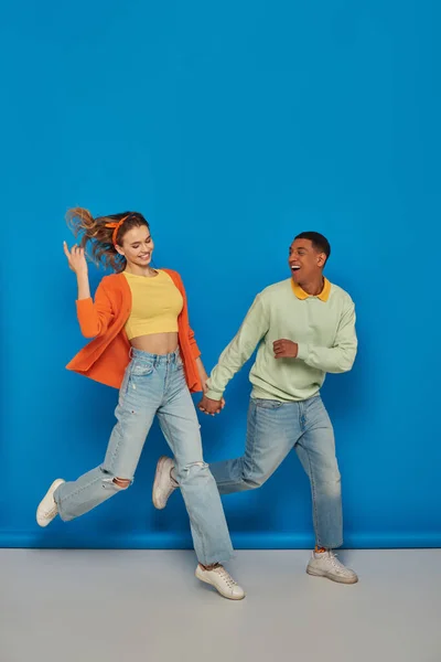 Fröhliches multikulturelles Paar in lässiger Kleidung, Händchen haltend und auf blauem Hintergrund laufend, lächelnd — Stockfoto