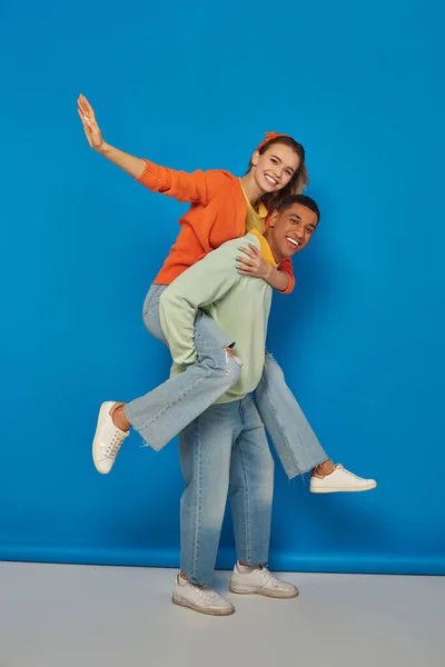 Feliz africano americano hombre piggybacking alegre novia en azul fondo, divertirse - foto de stock