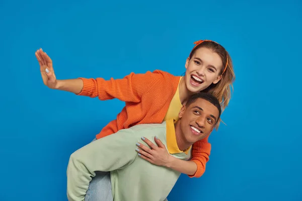 Joyeux afro-américain homme piggybackking petite amie joyeuse sur fond bleu, couple s'amuser — Photo de stock