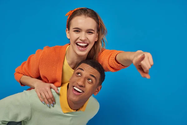 Fröhliche afrikanisch-amerikanische Mann huckepack fröhliche Freundin auf blauem Hintergrund, zeigt in die Kamera — Stockfoto